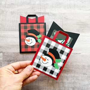May include: Two red and black plaid gift card holders with snowman faces. The holders are folded to create a pocket for a gift card.