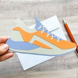 May include: A papercraft model of a blue and orange running shoe with white laces. The shoe is sitting on a white envelope.