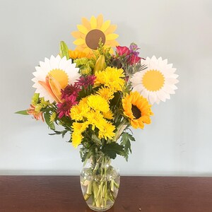 May include: A bouquet of yellow, white, and pink flowers in a clear glass vase. The flowers include sunflowers, daisies, and chrysanthemums.