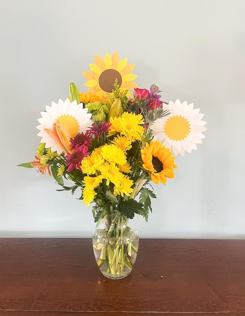 May include: A bouquet of yellow, white, and pink flowers in a clear glass vase. The flowers include sunflowers, daisies, and chrysanthemums.