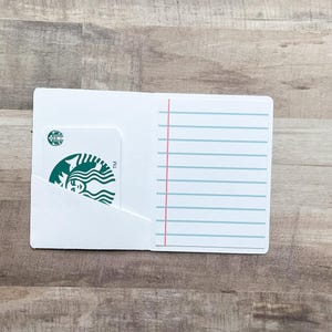 May include: White card holder with a Starbucks gift card partially visible in a pocket. The card holder has lined paper on the right side with a pink vertical line and blue horizontal lines. The background is a wooden surface.