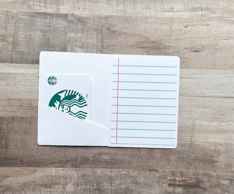 May include: White card holder with a Starbucks gift card partially visible in a pocket. The card holder has lined paper on the right side with a pink vertical line and blue horizontal lines. The background is a wooden surface.