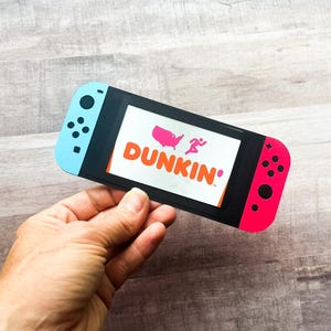 May include: A paper cutout of a video game controller with blue and red sides. The screen displays the Dunkin' logo with a pink map of the United States and a running figure. The controller is held in a hand.
