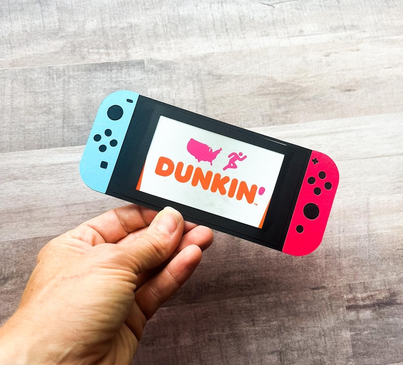 Peut inclure: D&eacute;coupe en papier d'une manette de jeu vid&eacute;o avec des c&ocirc;t&eacute;s bleus et rouges. L'&eacute;cran affiche le logo Dunkin' avec une carte rose des &Eacute;tats-Unis et une silhouette en course. La manette est tenue dans une main.