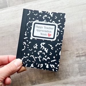 May include: A small notebook with a black and white speckled cover is held in a hand. The notebook has a black spine and a white label that reads "Happy Teacher Appreciation Week" with a small red heart.