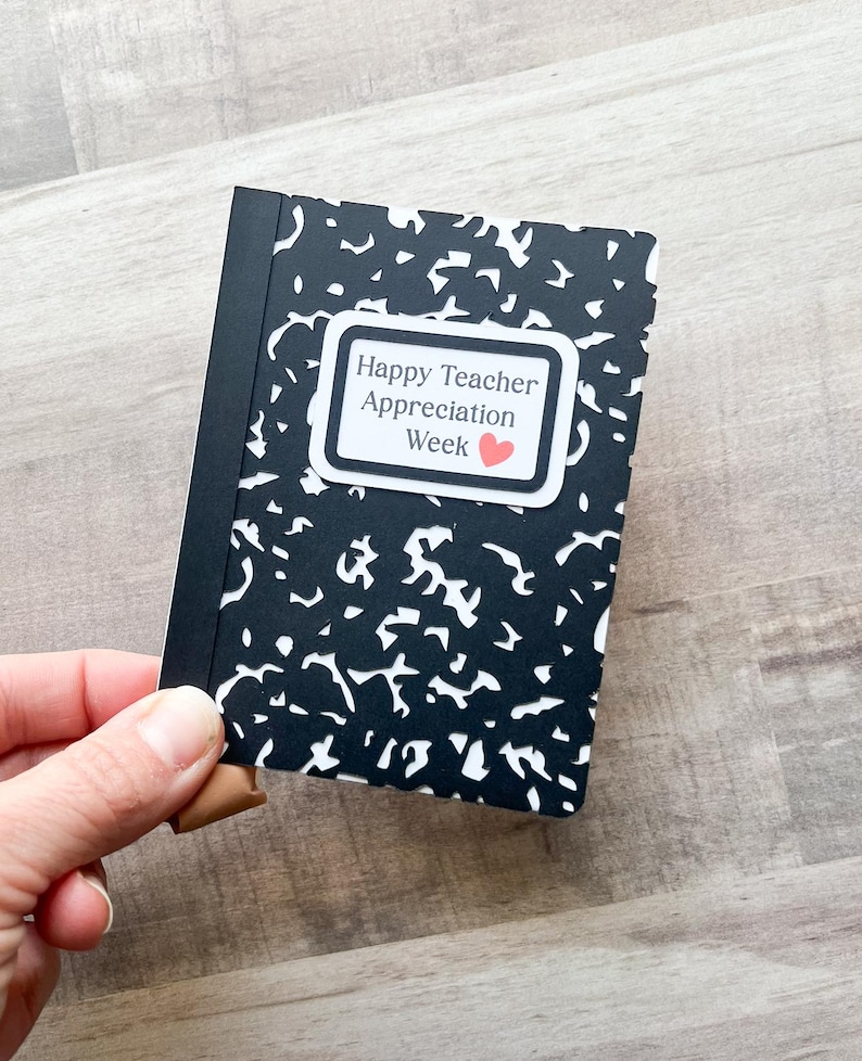 May include: A small notebook with a black and white speckled cover is held in a hand. The notebook has a black spine and a white label that reads "Happy Teacher Appreciation Week" with a small red heart.