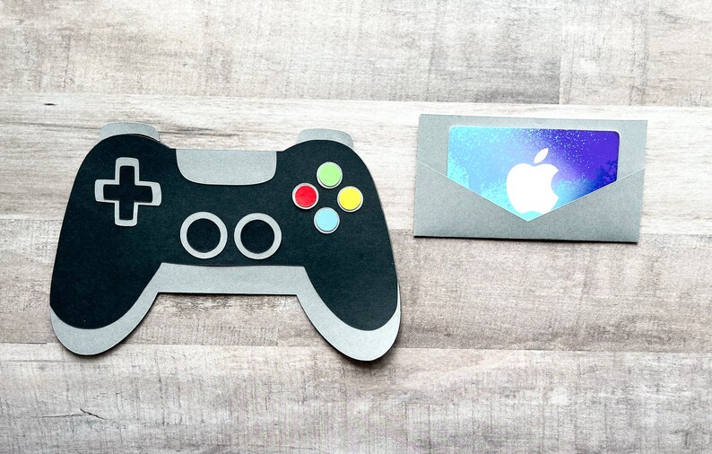 May include: A black and gray video game controller with colorful buttons next to a gray envelope with a blue and purple gift card featuring the Apple logo.