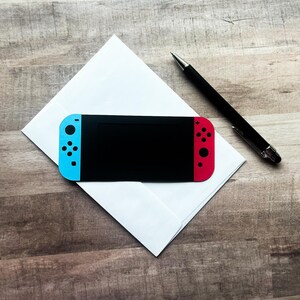 Peut inclure: Une console de jeu portable noire avec des contr&ocirc;leurs bleus et rouges est pos&eacute;e sur une enveloppe blanche. Un stylo noir est plac&eacute; &agrave; proximit&eacute; sur une surface en bois. La console a un &eacute;cran noir.