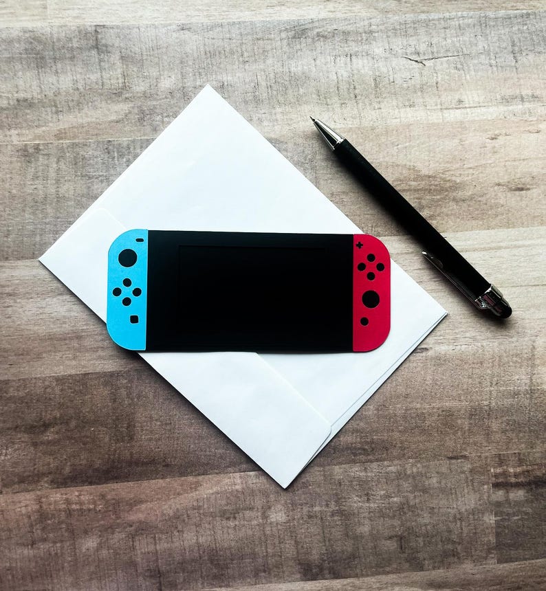 Peut inclure: Une console de jeu portable noire avec des contr&ocirc;leurs bleus et rouges est pos&eacute;e sur une enveloppe blanche. Un stylo noir est plac&eacute; &agrave; proximit&eacute; sur une surface en bois. La console a un &eacute;cran noir.