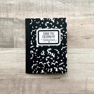 May include: A small, black and white composition notebook with a unique pattern. The notebook features a white rectangular label with the text "THANK YOU FOR BEING MY TEACHER" in black script. A heart is below the text.