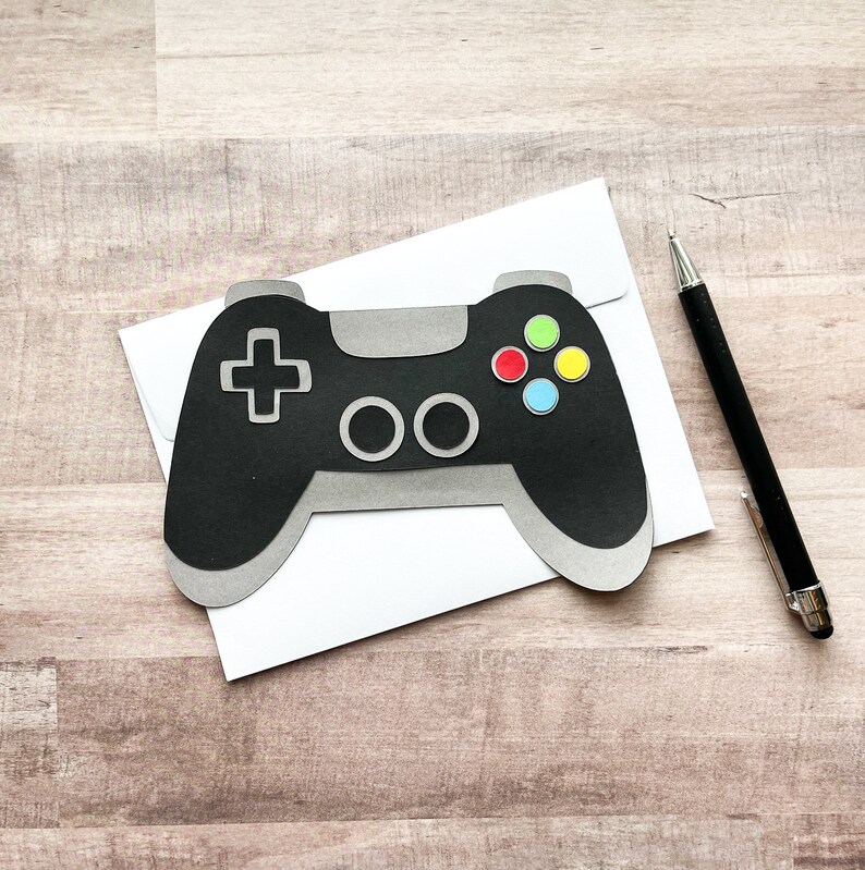 May include: A black and gray paper craft of a video game controller with colorful buttons on a white envelope.