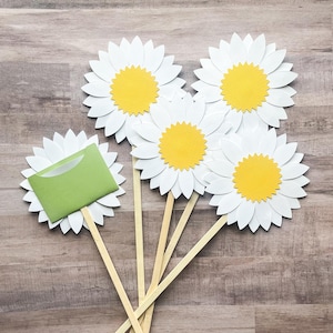 May include: Five white paper daisy flowers with yellow centers on wooden sticks. The flowers are arranged in a fan shape. One flower has a green envelope attached to it.