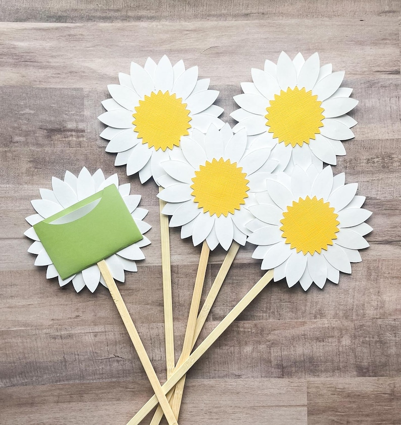 May include: Five white paper daisy flowers with yellow centers on wooden sticks. The flowers are arranged in a fan shape. One flower has a green envelope attached to it.