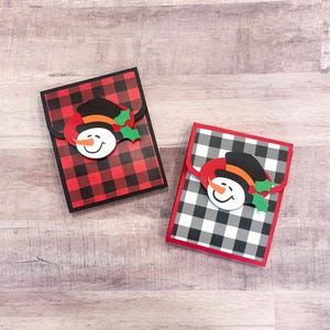 May include: Two decorative gift card holders with a snowman design. One has a red and black buffalo plaid pattern, the other has a black and white buffalo plaid pattern. Each has a snowman face with a black hat, orange scarf, and holly.