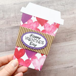 May include: A paper coffee cup with a brown cardboard sleeve and a purple heart-shaped label that says "Happy Valentine's Day". The cup is decorated with pink and purple watercolor hearts.