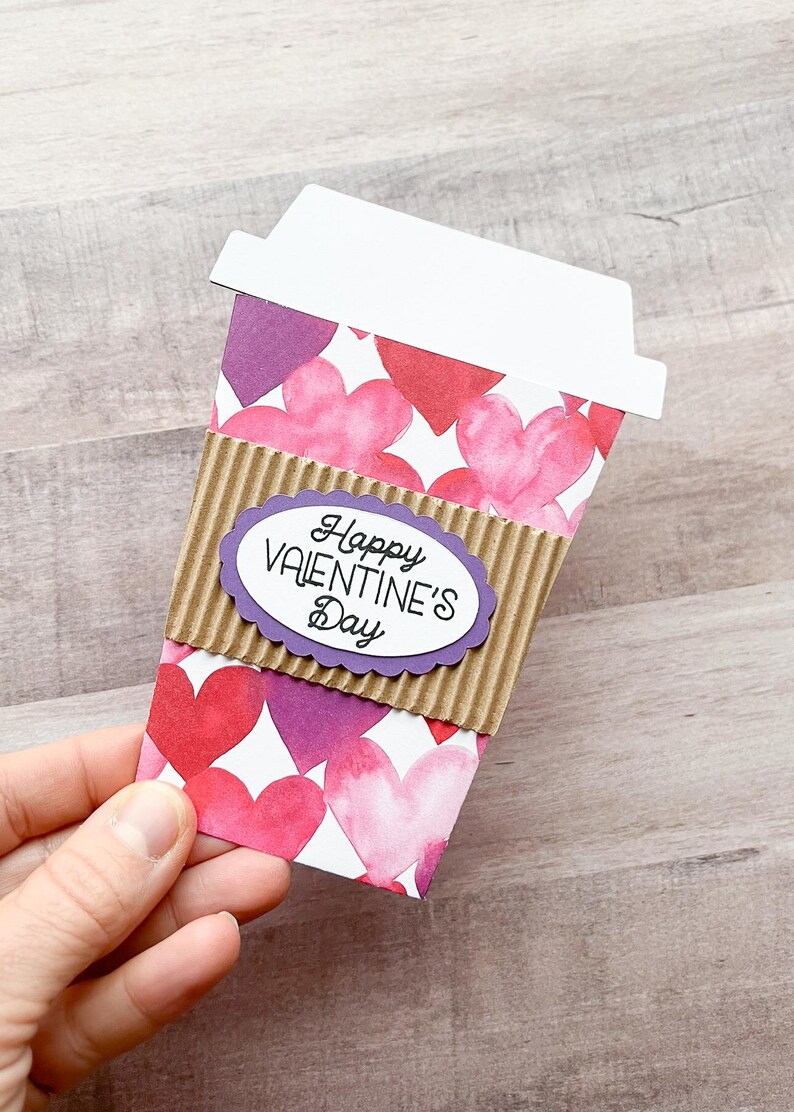 May include: A paper coffee cup with a brown cardboard sleeve and a purple heart-shaped label that says "Happy Valentine's Day". The cup is decorated with pink and purple watercolor hearts.