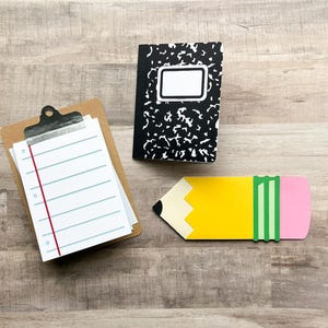 May include: A composition notebook with a black and white cover, a clipboard with lined paper, and a yellow pencil-shaped object with a pink eraser and green stripes. The items are arranged on a wooden surface.