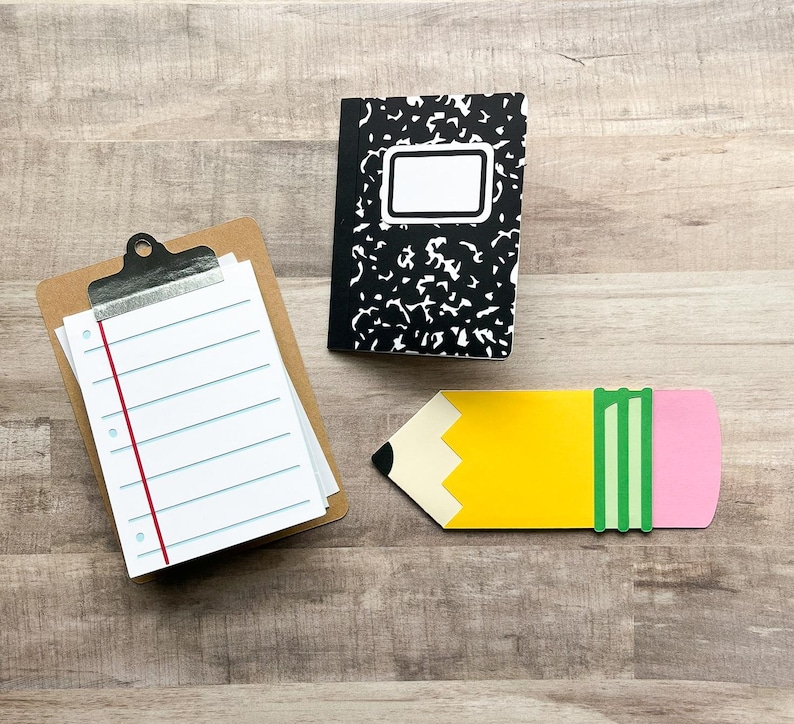 May include: A composition notebook with a black and white cover, a clipboard with lined paper, and a yellow pencil-shaped object with a pink eraser and green stripes. The items are arranged on a wooden surface.