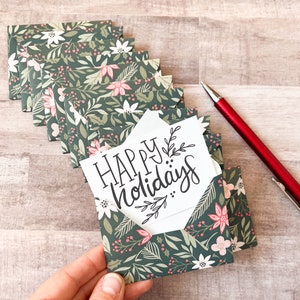 May include: A stack of holiday cards with a floral pattern in green, white, and pink. Each card is in a small envelope. One card is visible, with the words "HAPPY holidays" in black script. A red pen rests nearby.