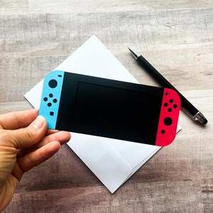 Peut inclure: Une carte noire en forme de console de jeu vid&eacute;o, avec des sections de manette bleues et rouges. Elle est tenue au-dessus d'une enveloppe blanche, avec un stylo noir &agrave; c&ocirc;t&eacute;. La console pr&eacute;sente un &eacute;cran noir et des d&eacute;tails de boutons.