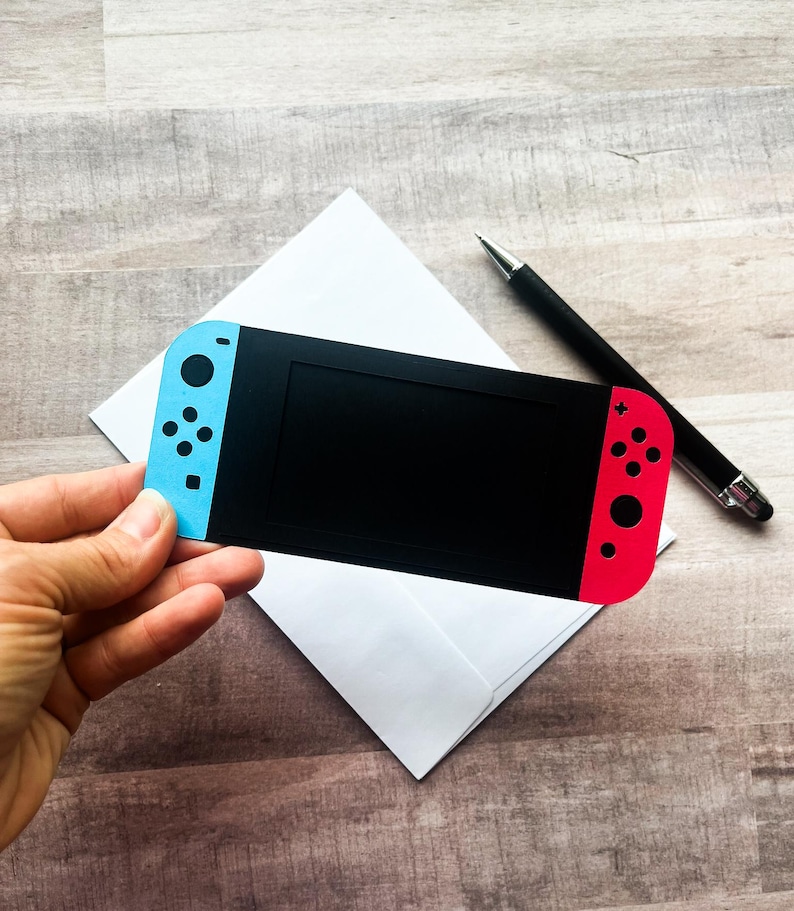Peut inclure: Une carte noire en forme de console de jeu vid&eacute;o, avec des sections de manette bleues et rouges. Elle est tenue au-dessus d'une enveloppe blanche, avec un stylo noir &agrave; c&ocirc;t&eacute;. La console pr&eacute;sente un &eacute;cran noir et des d&eacute;tails de boutons.