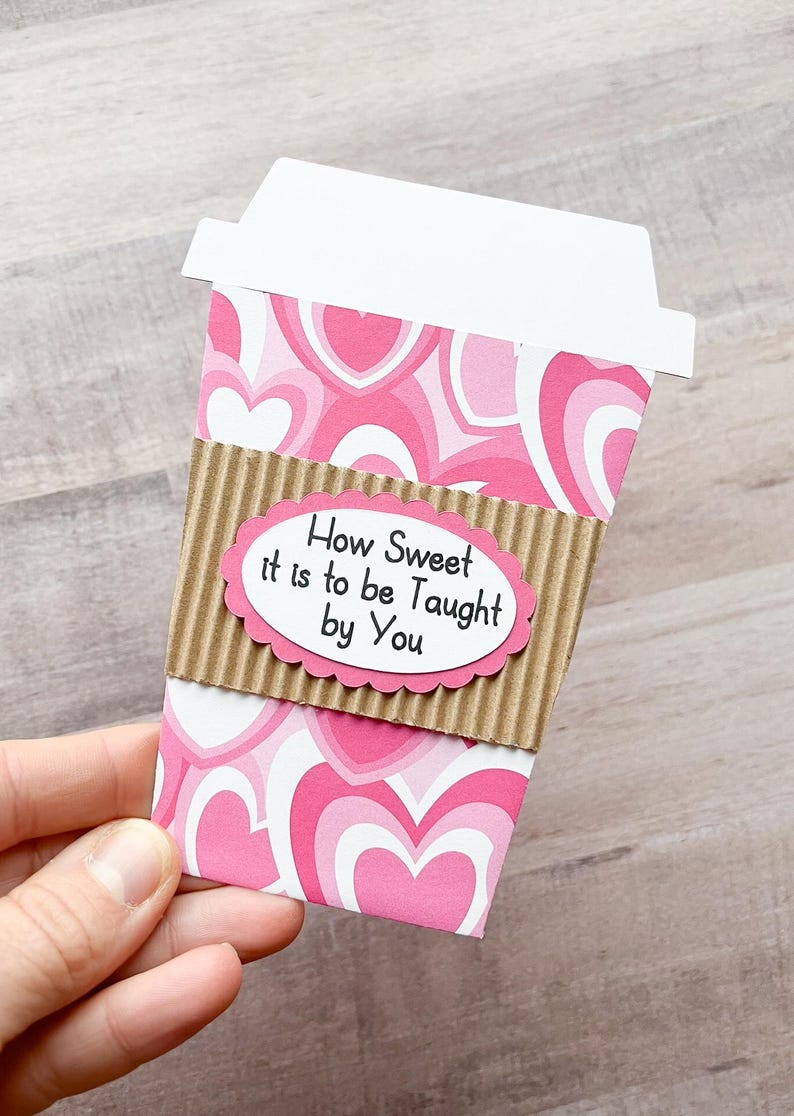 May include: A pink and white coffee cup with a brown cardboard sleeve. The sleeve has a white oval sticker with the text "How Sweet it is to be Taught by You".