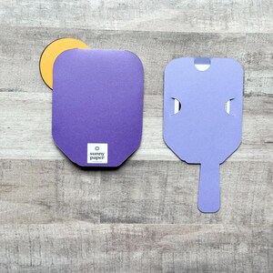 May include: Two purple cardboard boxes with a cut-out handle. The box on the left has a round yellow sticker with the text "sunny paper studio" on it. The box on the right has a cut-out handle in the shape of a popsicle.