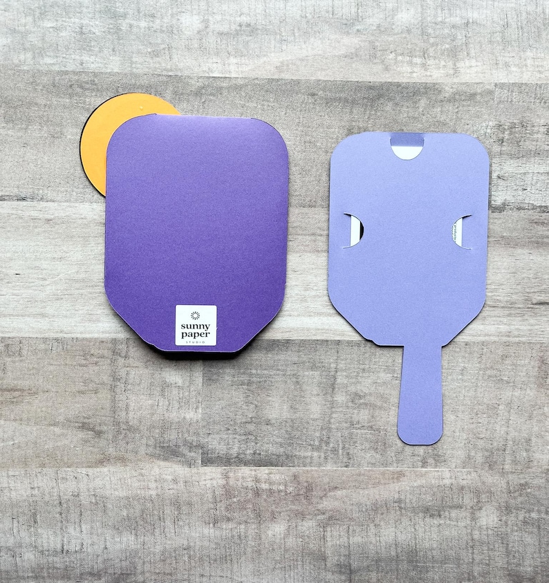 May include: Two purple cardboard boxes with a cut-out handle. The box on the left has a round yellow sticker with the text "sunny paper studio" on it. The box on the right has a cut-out handle in the shape of a popsicle.