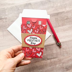 May include: A red and brown paper coffee cup gift card holder with a white lid. The cup is decorated with pink hearts and the text "Happy Valentine's Day".