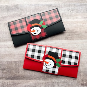 May include: Two red and black plaid gift card holders with a snowman design. The snowman has a black hat, orange nose, and green holly.