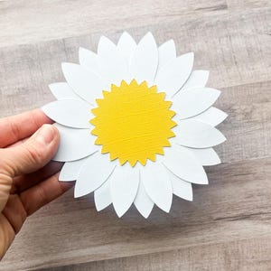 May include: A white paper daisy with a yellow center. The daisy is made of multiple layers of paper, giving it a 3D effect.