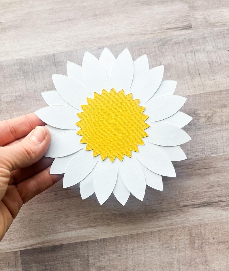 May include: A white paper daisy with a yellow center. The daisy is made of multiple layers of paper, giving it a 3D effect.