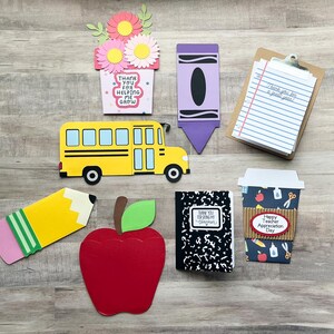 May include: A collection of colorful, die-cut paper crafts with a school theme. Includes a yellow school bus, a red apple, a pencil, a crayon, a notepad, a coffee cup, a notebook, and a flower pot with the text "Thank you for helping me grow."