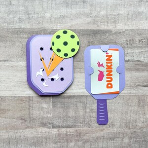 May include: Two purple paper crafts shaped like a pickleball paddle. One paddle has a green pickleball on top and the other has a Dunkin' gift card inside.