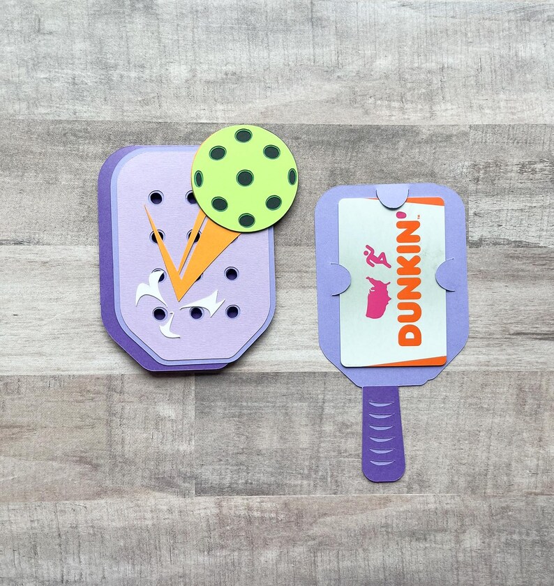 May include: Two purple paper crafts shaped like a pickleball paddle. One paddle has a green pickleball on top and the other has a Dunkin' gift card inside.