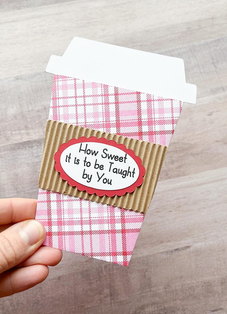 May include: A pink and white plaid coffee cup with a brown cardboard sleeve. The sleeve has a red oval with the text "How Sweet it is to be Taught by You".