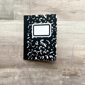 May include: A composition notebook with a black and white speckled cover design. The notebook has a white rectangular label on the front cover. The spine is black.