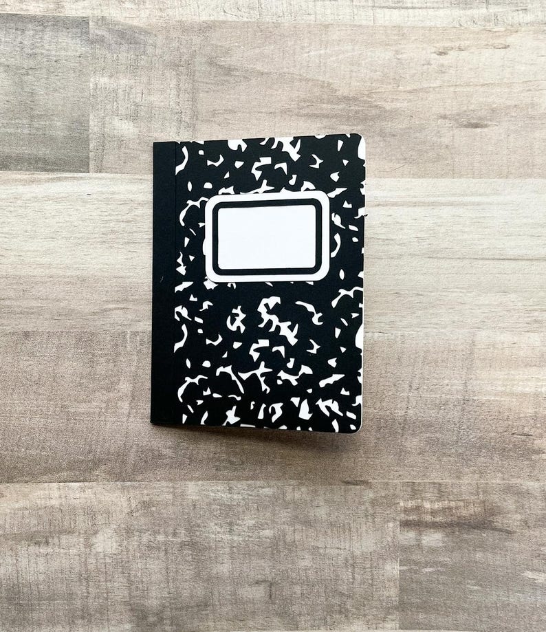 May include: A composition notebook with a black and white speckled cover design. The notebook has a white rectangular label on the front cover. The spine is black.