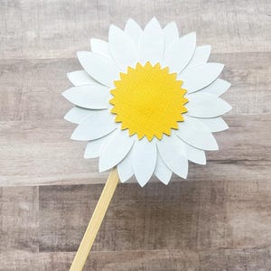 May include: A white paper daisy with a yellow center on a wooden stick. The daisy is a craft supply for a party or event.