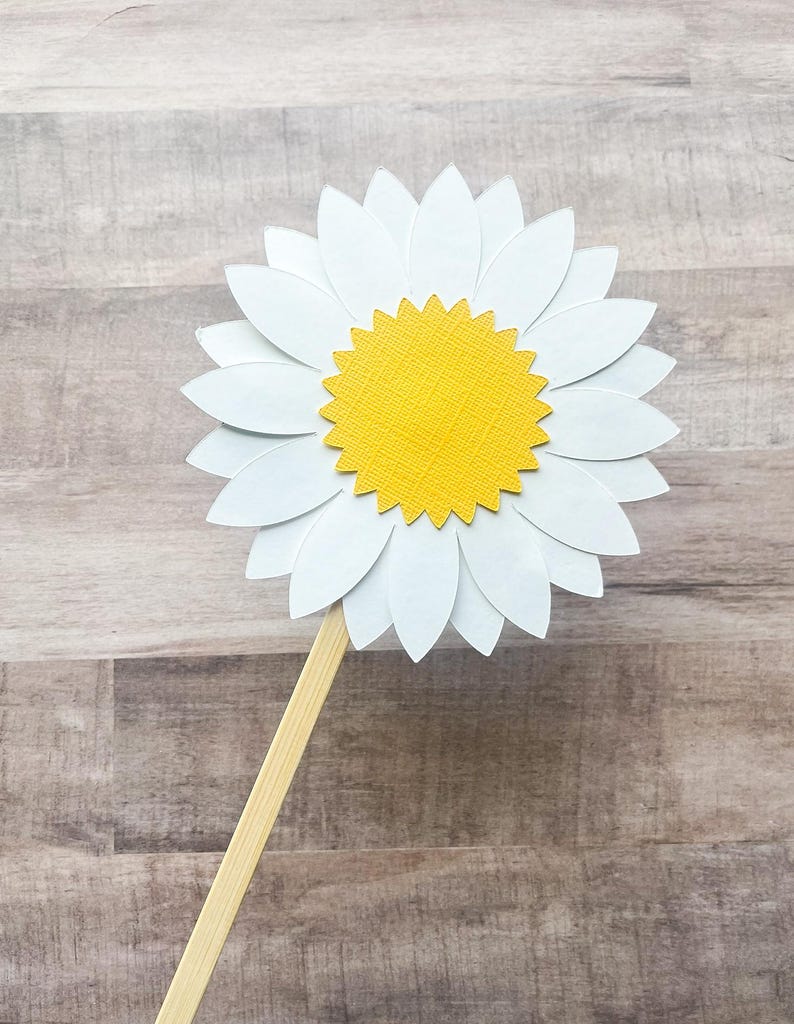May include: A white paper daisy with a yellow center on a wooden stick. The daisy is a craft supply for a party or event.