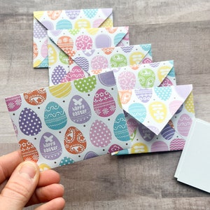 May include: A stack of colorful Easter-themed envelopes with a white background and a pattern of pastel-colored eggs. The top envelope has the text "Happy Easter" printed on it.