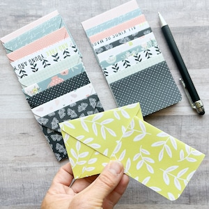 May include: A set of colorful envelopes with various patterns and text, including "And Free", "Young And", and "All I Want Is Magic". The envelopes are stacked on a light wood surface. A hand is holding a yellow envelope with a white leaf pattern.