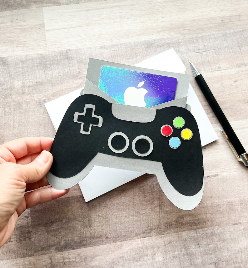 May include: A black and gray paper craft of a video game controller with a gift card tucked inside. The gift card is a blue and purple holographic design with the Apple logo.