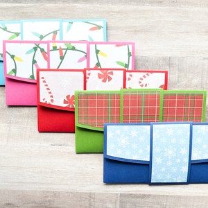 May include: Six Christmas card envelopes with different holiday designs. The envelopes are folded and stacked on a woodgrain surface. The designs include a red and green plaid, snowflakes, candy canes, and Christmas lights.