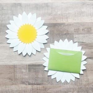 May include: Two white paper daisy flowers with yellow centers. One daisy has a green envelope inside.