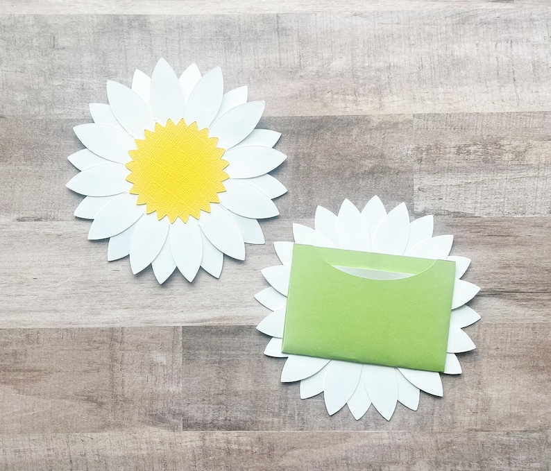 May include: Two white paper daisy flowers with yellow centers. One daisy has a green envelope inside.