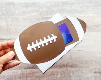 May include: A brown and white paper football-shaped gift card holder featuring white laces. The holder is designed to slide over a rectangular gift card with a blue and purple galaxy-themed pattern, resting on a plain white envelope.