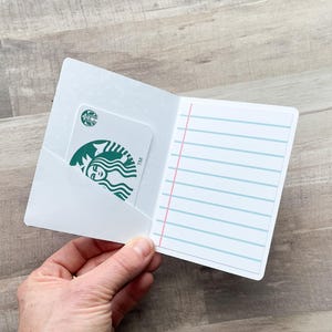 May include: A white card holder with a Starbucks gift card in the pocket. The gift card has the Starbucks logo. The right side of the card holder has blue lined paper with a red margin. The card holder is open.