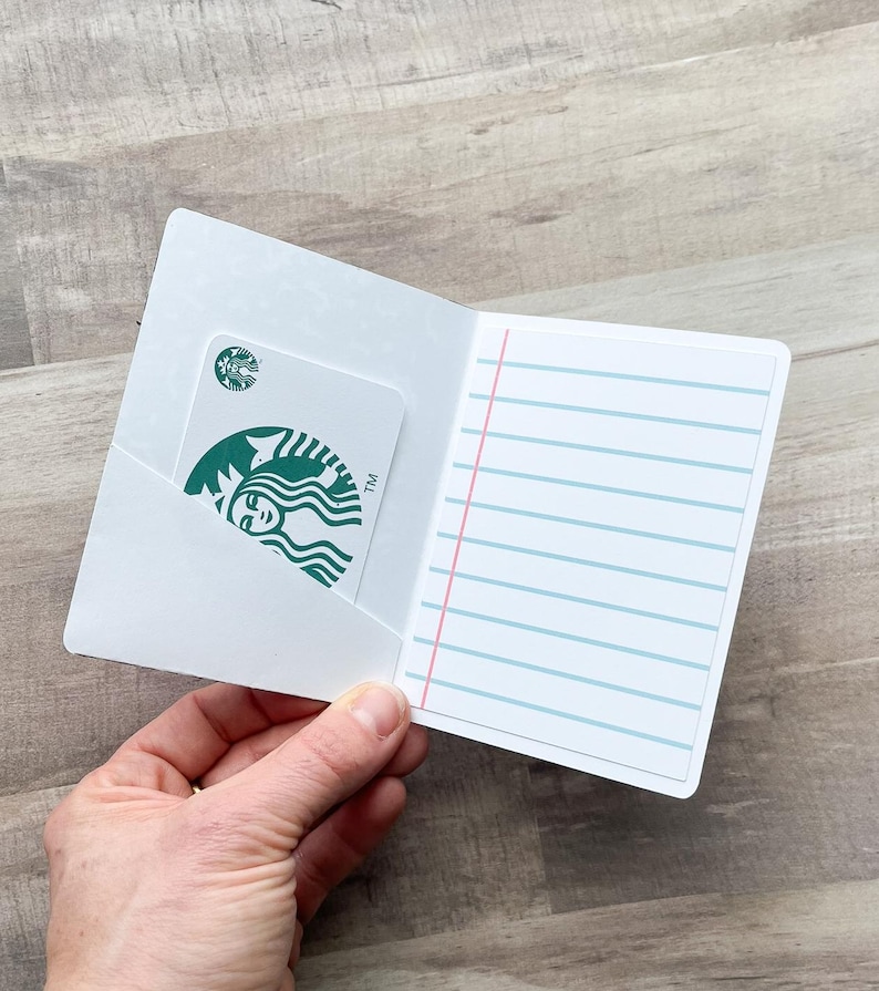 May include: A white card holder with a Starbucks gift card in the pocket. The gift card has the Starbucks logo. The right side of the card holder has blue lined paper with a red margin. The card holder is open.