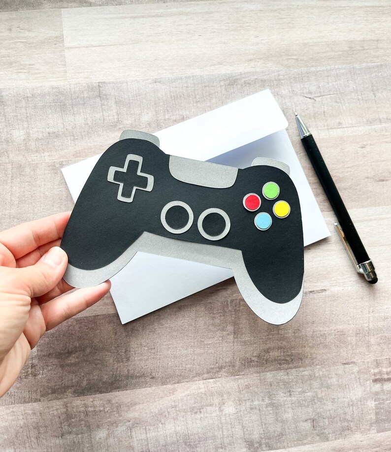 May include: A black and silver video game controller with colorful buttons. The controller is on a white card with a white envelope.